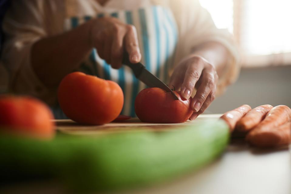 Person slicing tomatoes on a wooden board, surrounded by various vegetables, in a kitchen setting