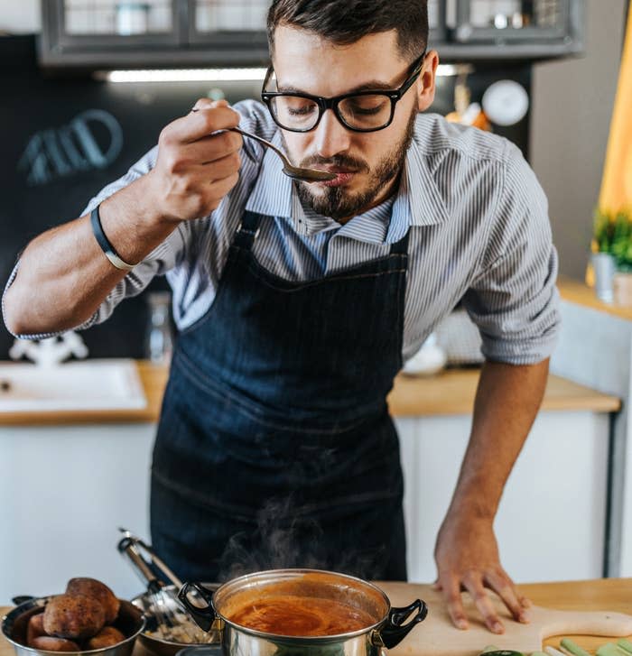 Man in a kitchen tasting soup from a pot, wearing glasses and an apron, surrounded by vegetables