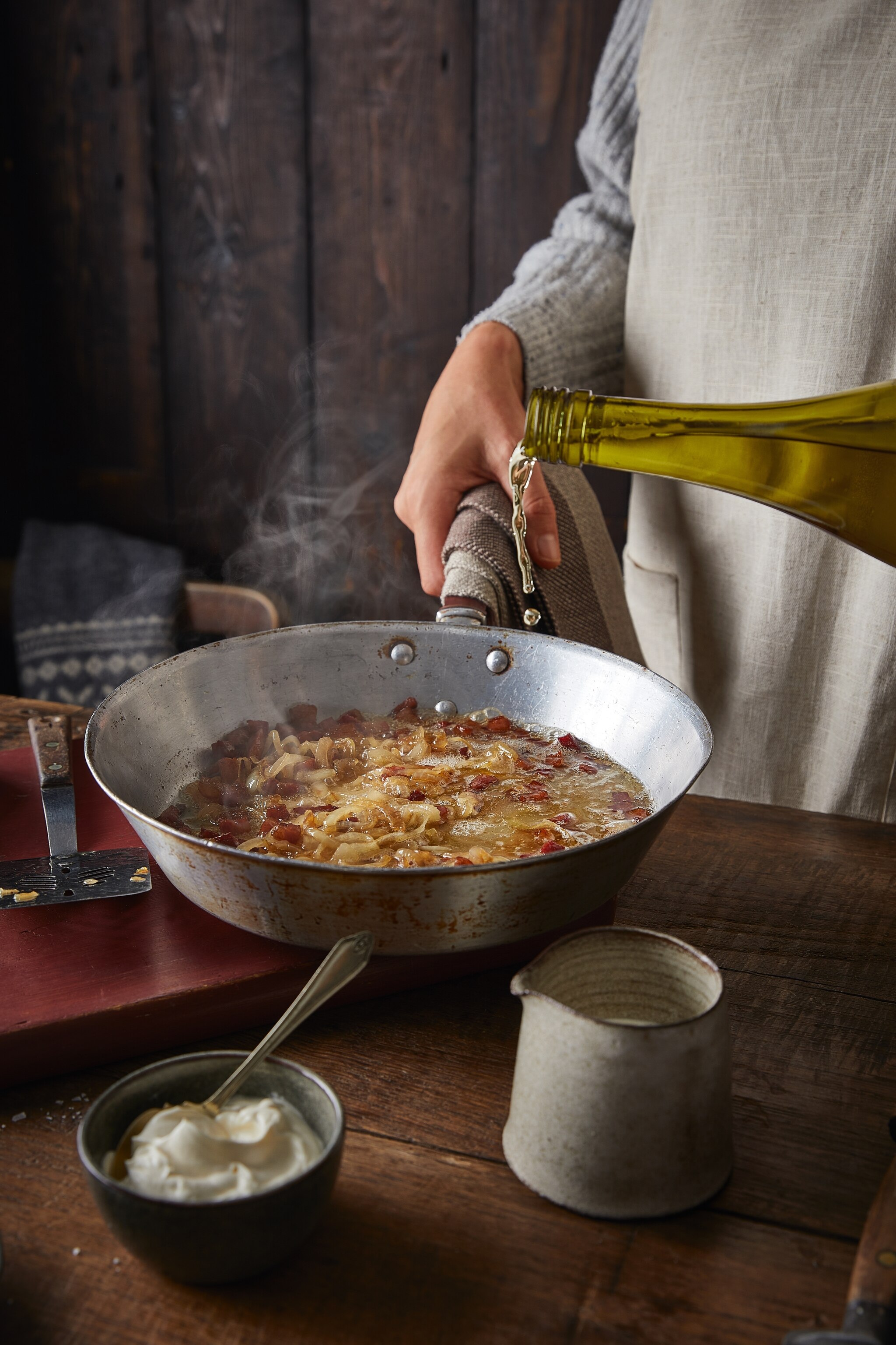 A chef adding wine to a sizzling frying pan of bacon and onions.