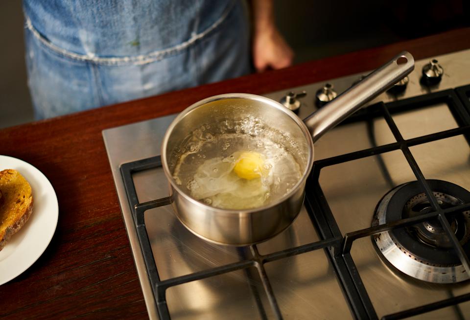 A pot of water on a stove with a poaching egg inside, next to a slice of toast on a plate