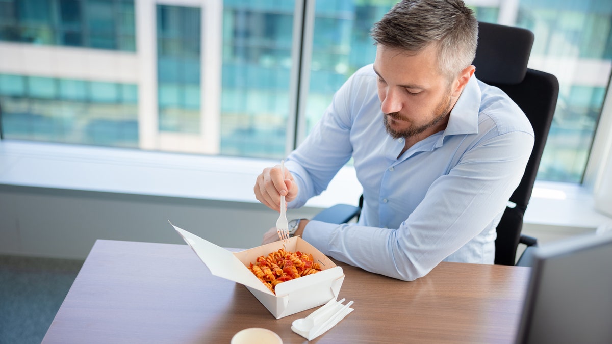 A man in a blue dress shirt eats pasta with a white plastic fork at his work desk.
