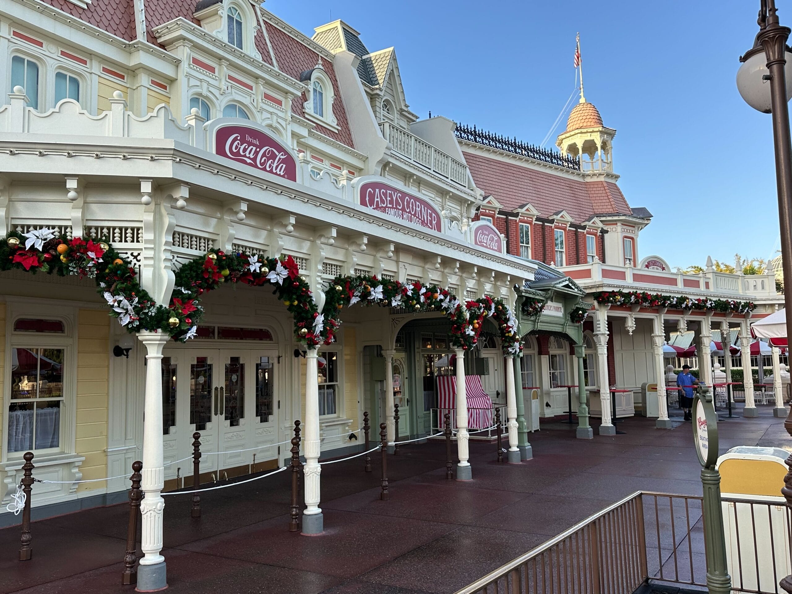 A Victorian-style building, decked with enchanting Christmas decorations, is illuminated with holiday wreaths and lights. It proudly displays a Coca-Cola sign and the name "Casey's Corner," capturing the festive spirit of Magic Kingdom.