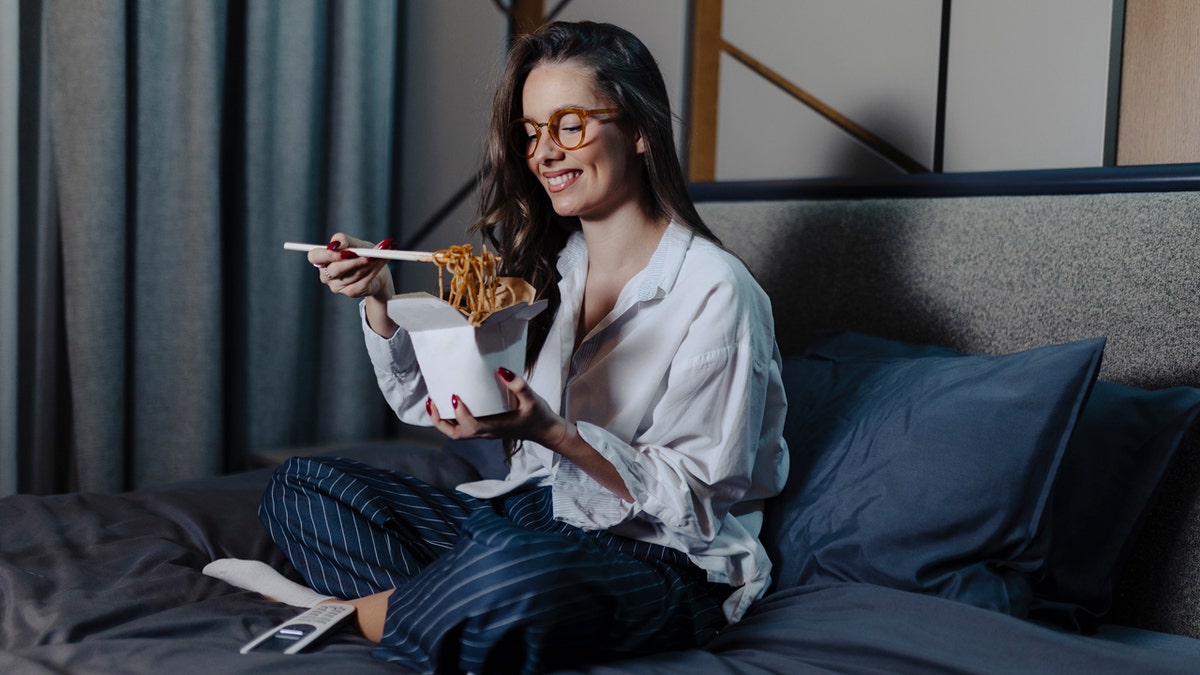 A woman with glasses smiles as she eats noodles with chopsticks in bed.