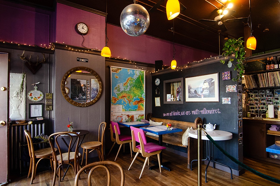 Interior of a cafe, featuring a disco ball, pink accents, a map of Europe, a big mirror with a decorative brass frame, string lights, and a quote painted in pink on a black wall: 