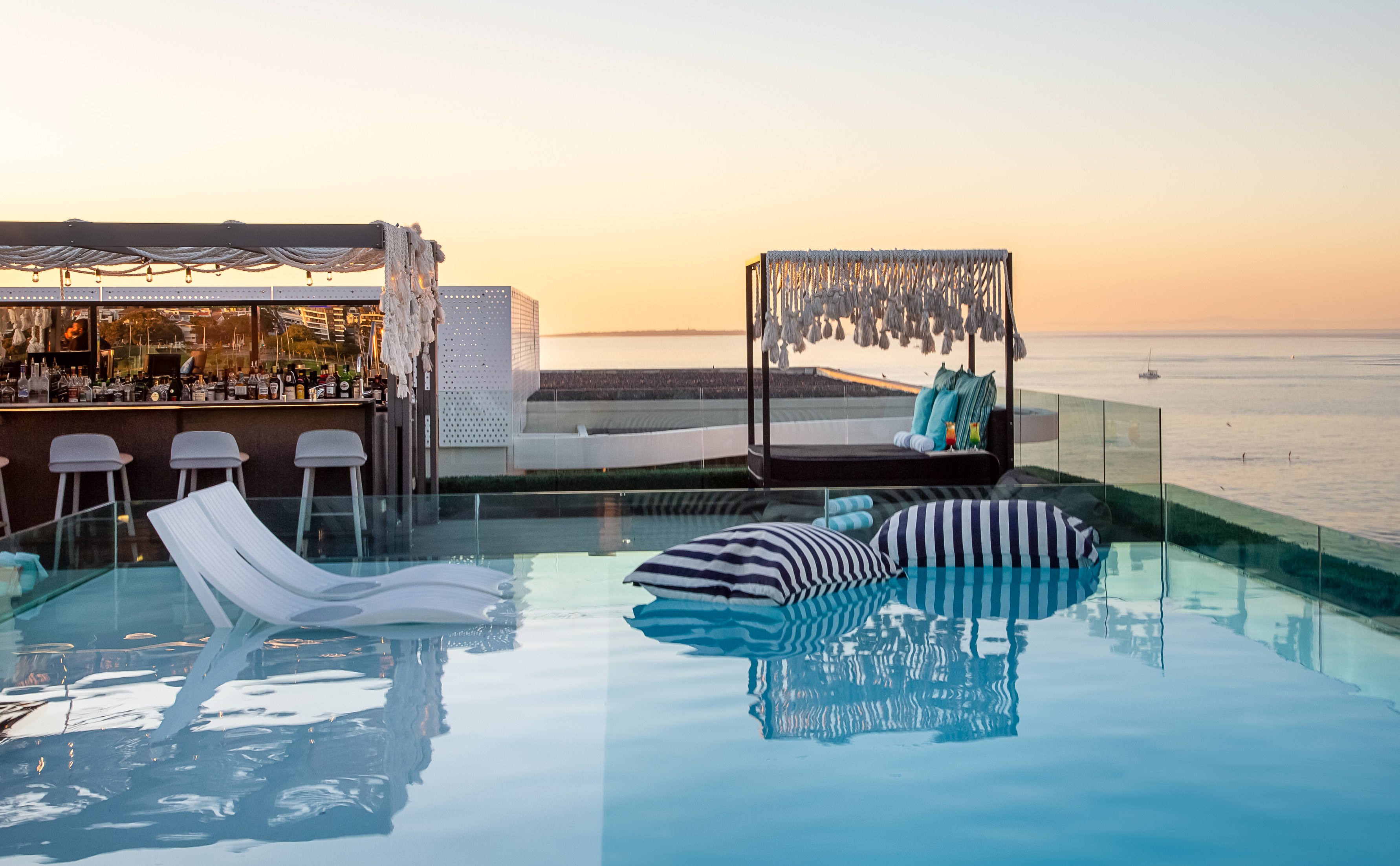 Rooftop infinity pool with bar and lounge area at sunset.