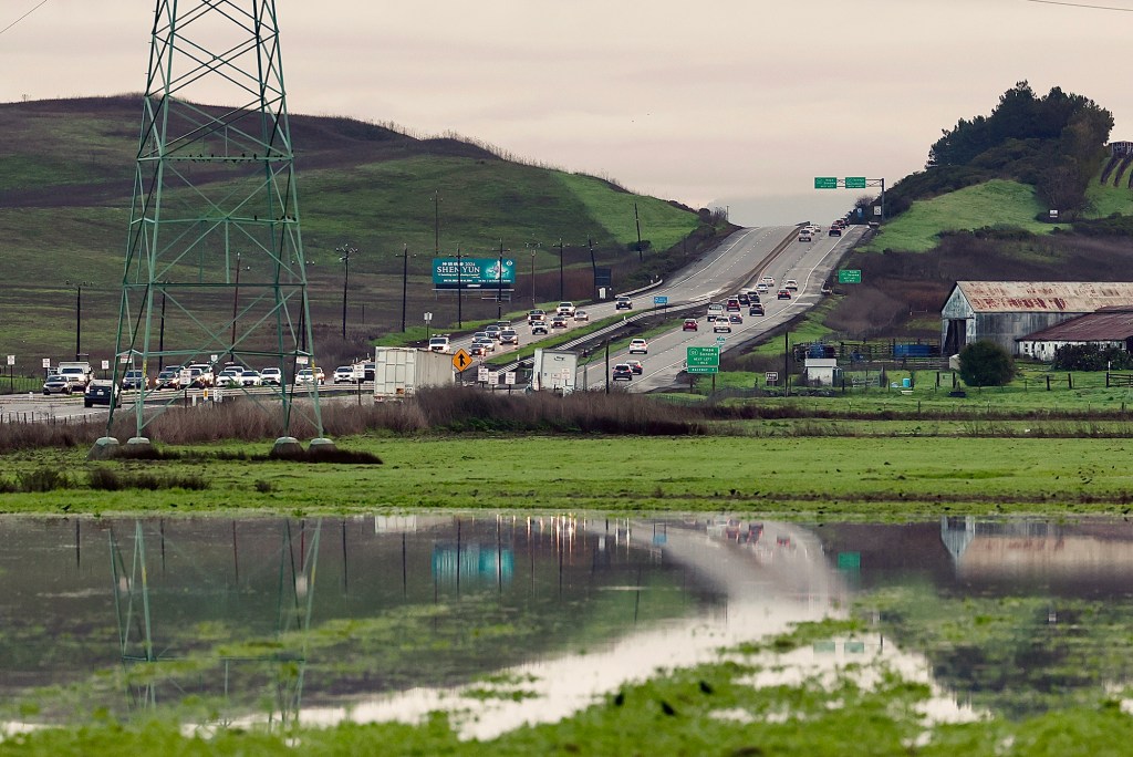 A stretch of Highway 37 in Napa County shows moderate traffic in both directions, surrounded by green hills and farmland. A large power line tower stands in the foreground near a flooded field that reflects the vehicles and landscape. Road signs and a billboard for Shen Yun are visible along the roadside.