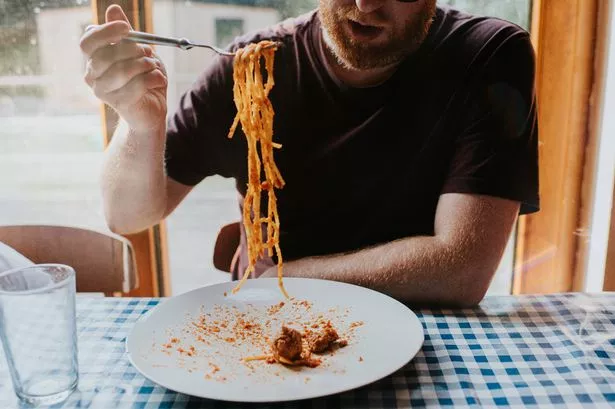 A man sits at a table, covered with a blue checkered tablecloth. He uses a fork to scoop up a mouthful of long spaghetti pasta. Meatballs rest on the large plate.