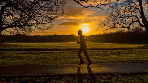 PA Media A jogger running in a park during sunrise, silhoutted against the rising sun as they run between trees
