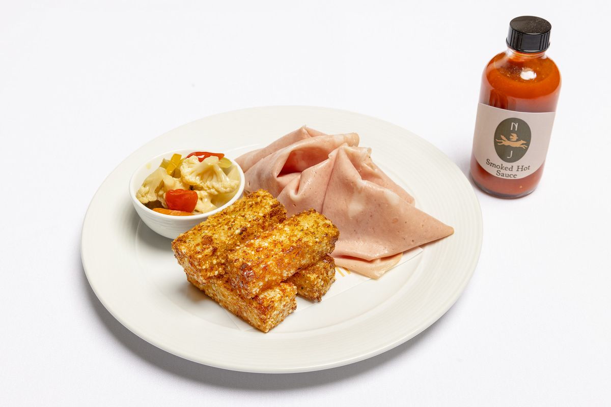 Slices of mortadella with pickles and four cheese fritters on a plate next to a bottle of red hot sauce.