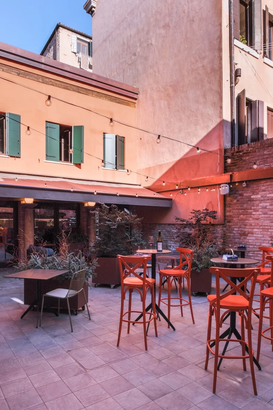 A tranquil piazza hosts a colorful wine bar whose tall, bright orange wooden stools and round wrought iron tables blend in naturally with the surrounding teracotta-hued buildings, dotted in fairylights.