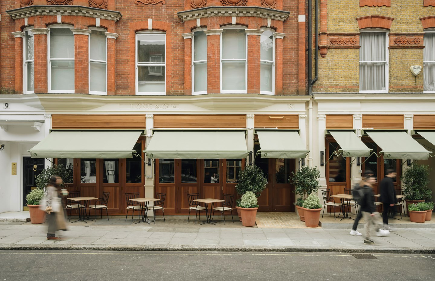 The exterior facade of a restaurant, sited within a historical red-brick townhouse, presents pale, muted green curtains and wood and glass finishings, as well as wrought iron garden table sets.