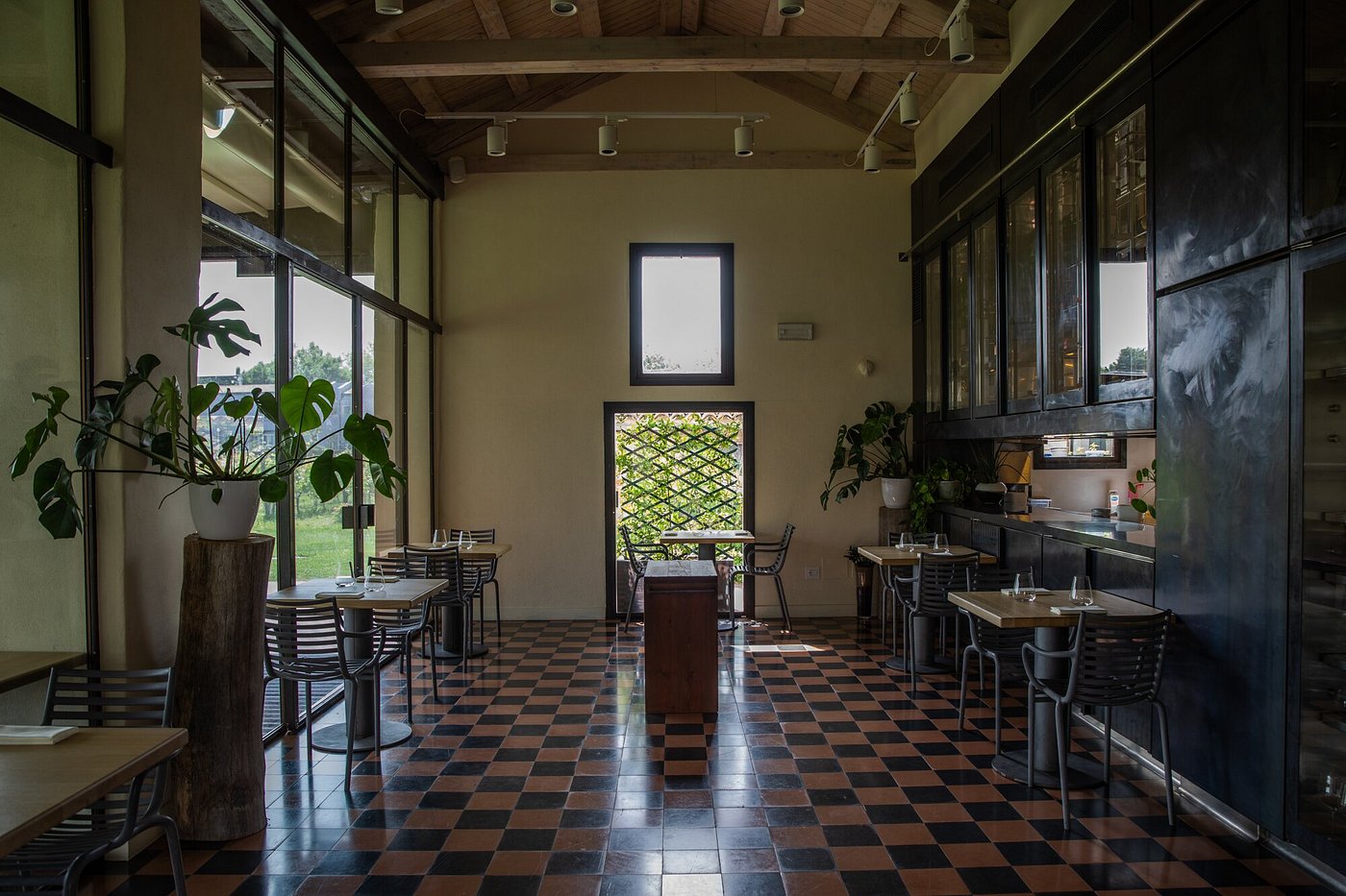 A countryside osteria features retro-inspired interiors with chequered salmon and green floors, industrial-style tables in wood with legs and chairs in wrought iron, trailing plants, yellow walls, and a relaxed, chic atmosphere.