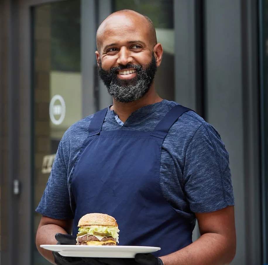 A smiling Taddesse holds up one of his classic burgers on a plate.