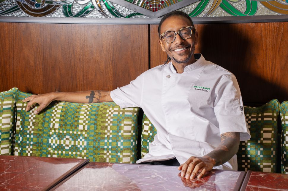 Chef Gregory Gourdet smiles while seated in a booth at Maison Passerelle, wearing his Printemps-branded chef’s jacket.