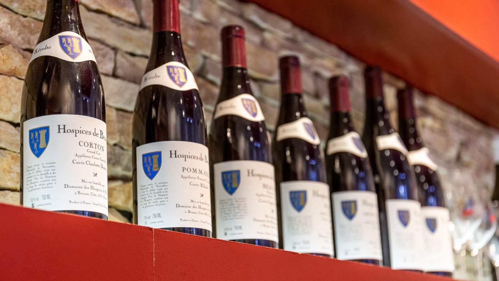 A close-up photograph featuring bottles of wine displayed on a red stand against a wall, bearing the inscription 'Hospices de Beaune.' The image focuses on the arrangement of the bottles, providing a detailed view of the wine labels and the overall presentation.