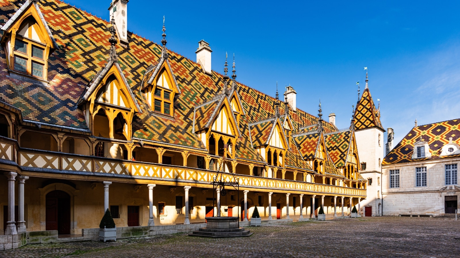 A photograph capturing the exterior of Hospices de Beaune, showcasing its architectural features. The image presents the historical building against a neutral sky, featuring characteristic elements such as pitched roofs and ornate windows.