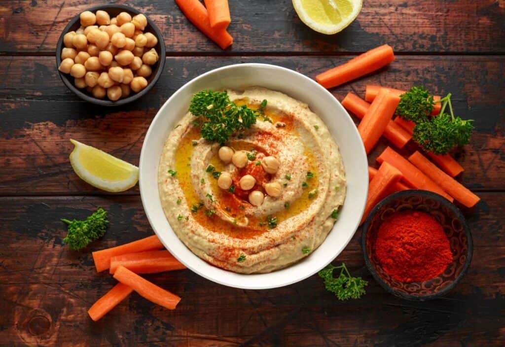 A bowl of hummus topped with chickpeas, parsley, olive oil, and paprika takes center stage for national hummus day, surrounded by carrot sticks, a bowl of chickpeas, lemon wedges, and a bowl of paprika on a wooden table.