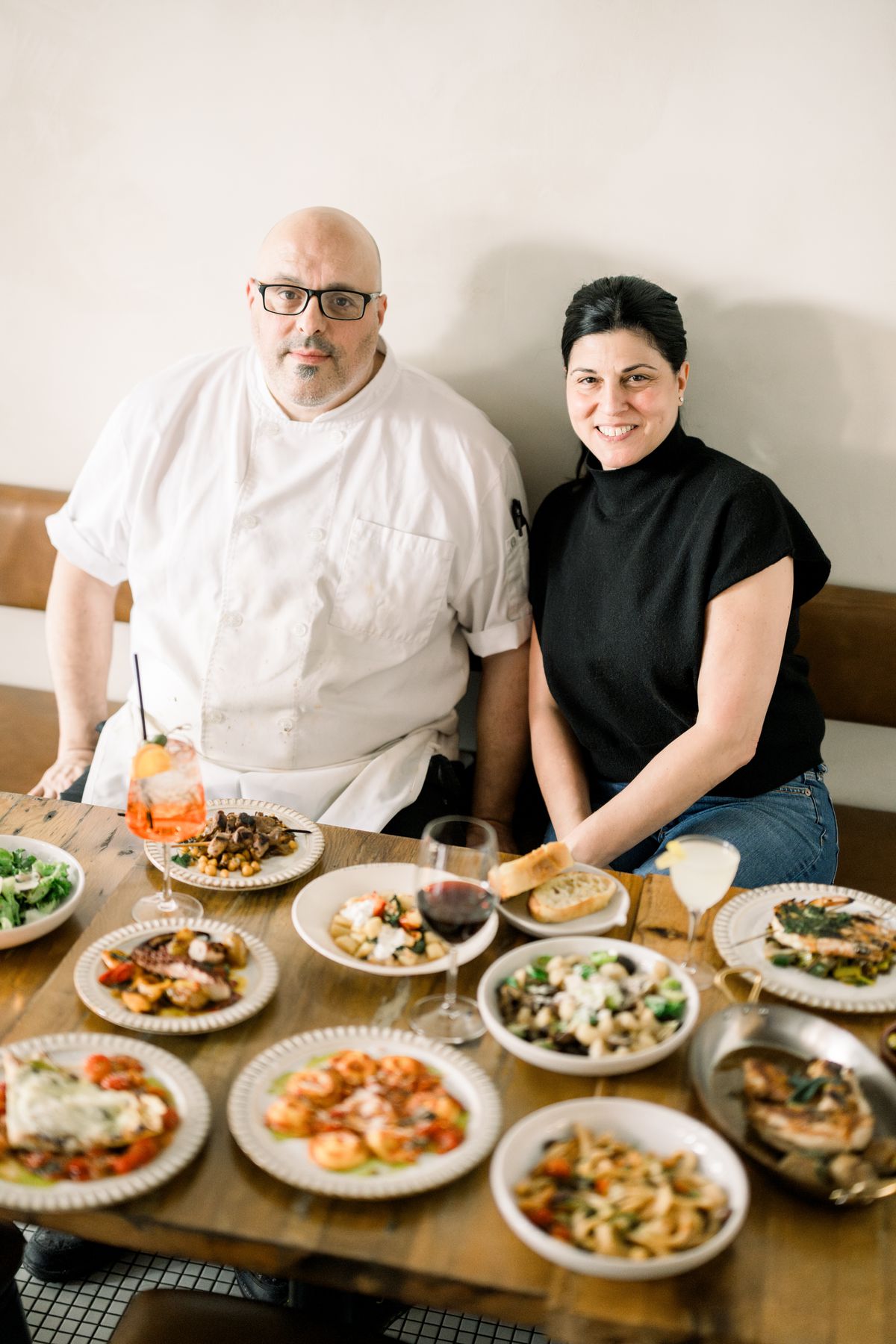 Two people sit smiling at the camera behind a restaurant table filled with food and drinks.