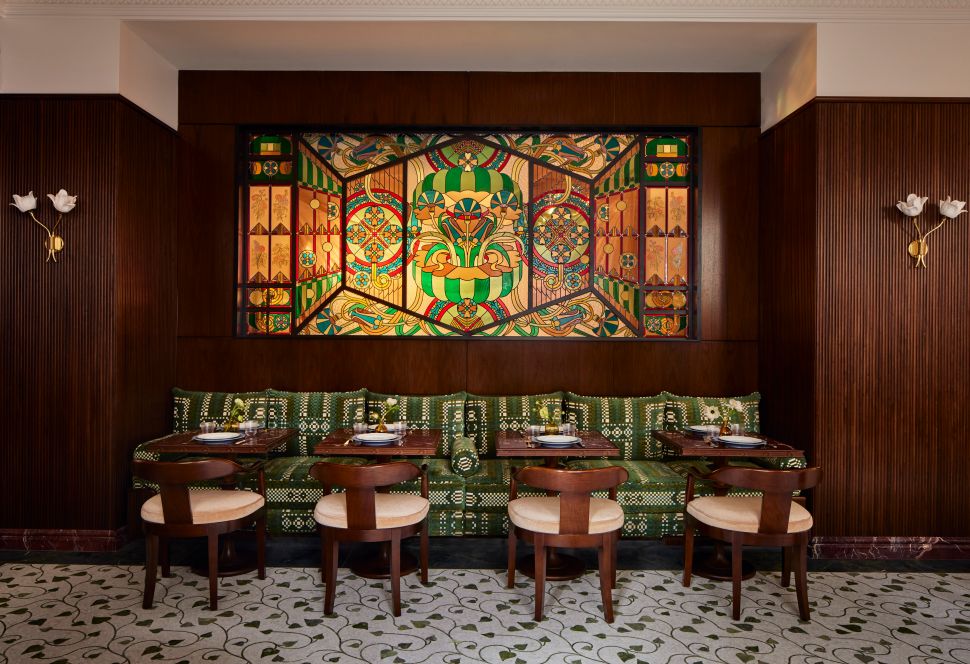 A row of tables and wood chairs line a green patterned banquette beneath a large, colorful stained glass mural at Maison Passerelle.