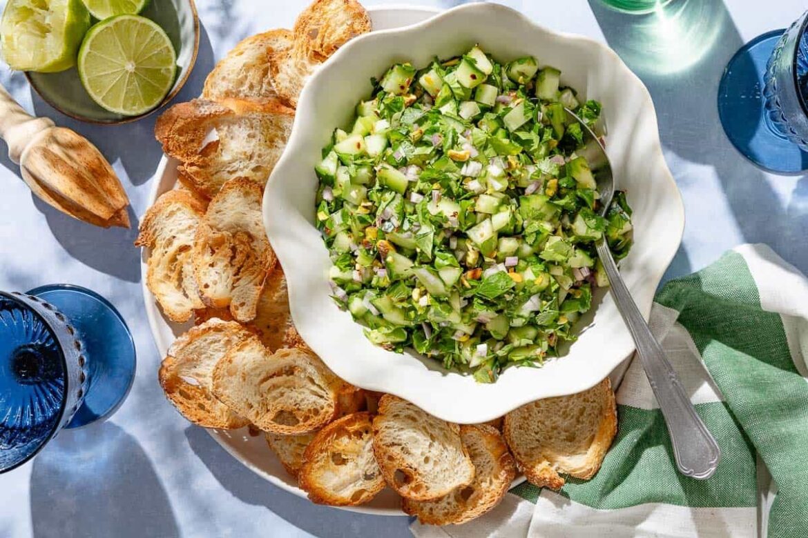 Cucumber Salsa | The Mediterranean Dish An overhead photo of cucumber salsa in a bowl with a spoon on a platter with slices of toasted baguette. Next to this is a kitchen towel, a plate of limes , a juicer and two glasses of water.