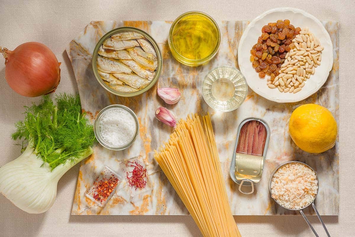 Ingredients for sardine pasta including sardines, spaghetti, onion, fennel, white wine, golden raisins, saffron, anchovy filets, garlic, red pepper flakes, lemon, pine nuts, bread crumbs and salt.