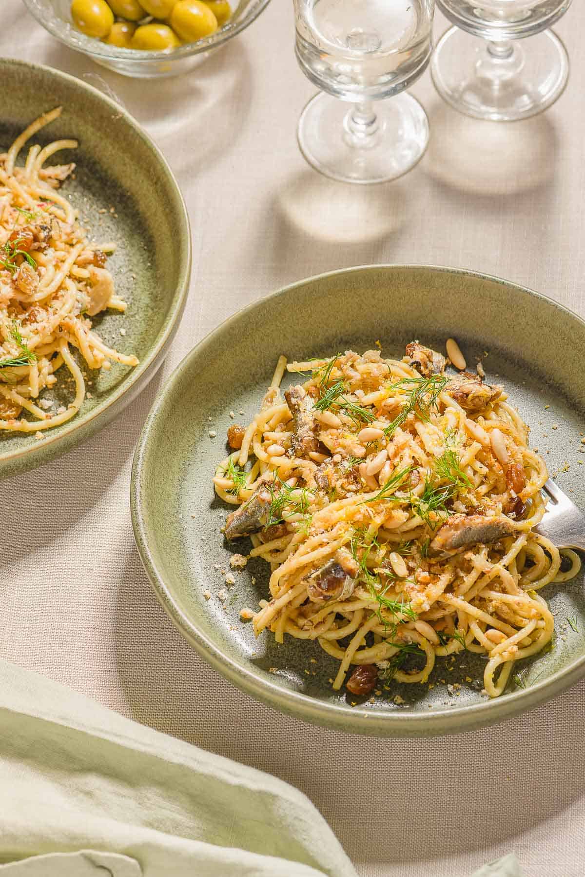 Two bowls of sardine pasta, one with a fork. These are surrounded by a bowl of green olives and two glasses of white wine.