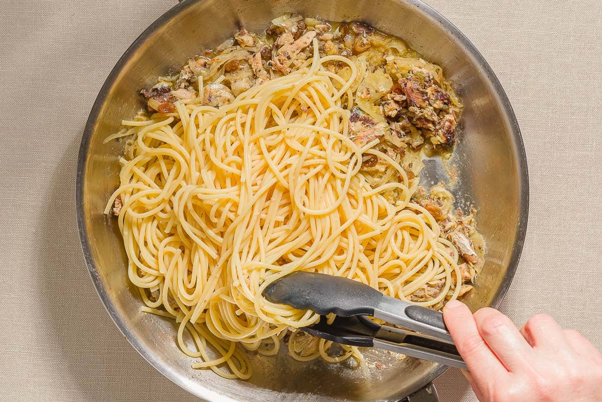 Cooked spaghetti being added to the sautéed sliced onions, fennel, sardines and anchovy fillets for the sardine pasta in a skillet.