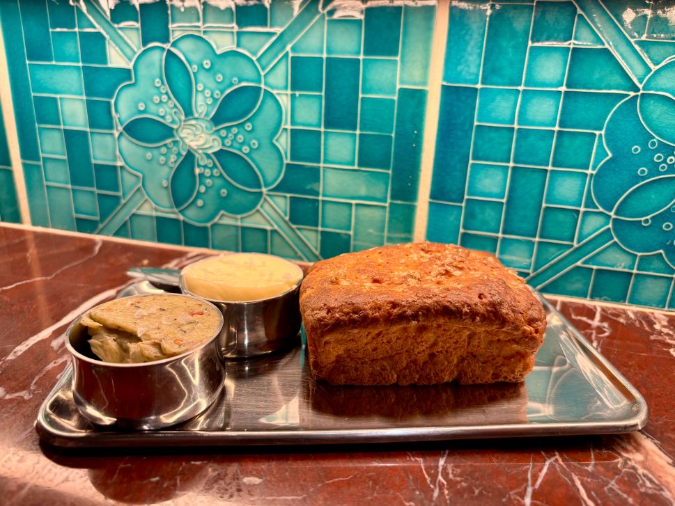 A loaf of warm plantain bread is served with two types of butter on a metal tray in front of turquoise tile.