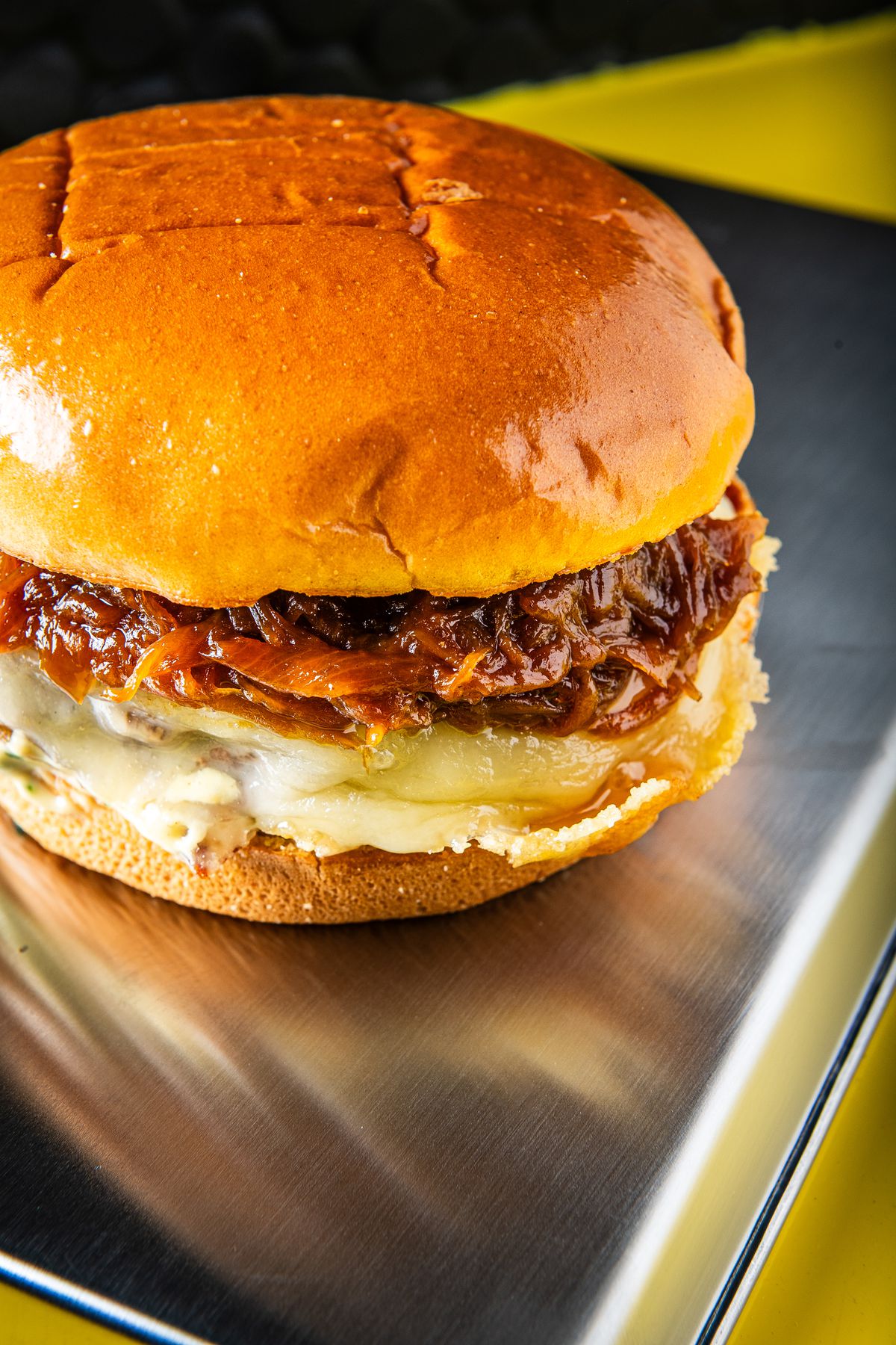 A burger with caramelized onion and melted cheese sits on a metallic counter.
