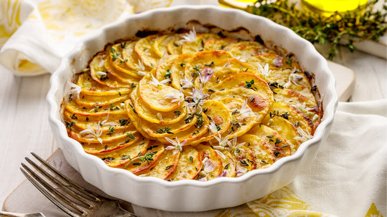 Scalloped potatoes in a white baking dish sit on a white and yellow cloth with a fork lying adjacent