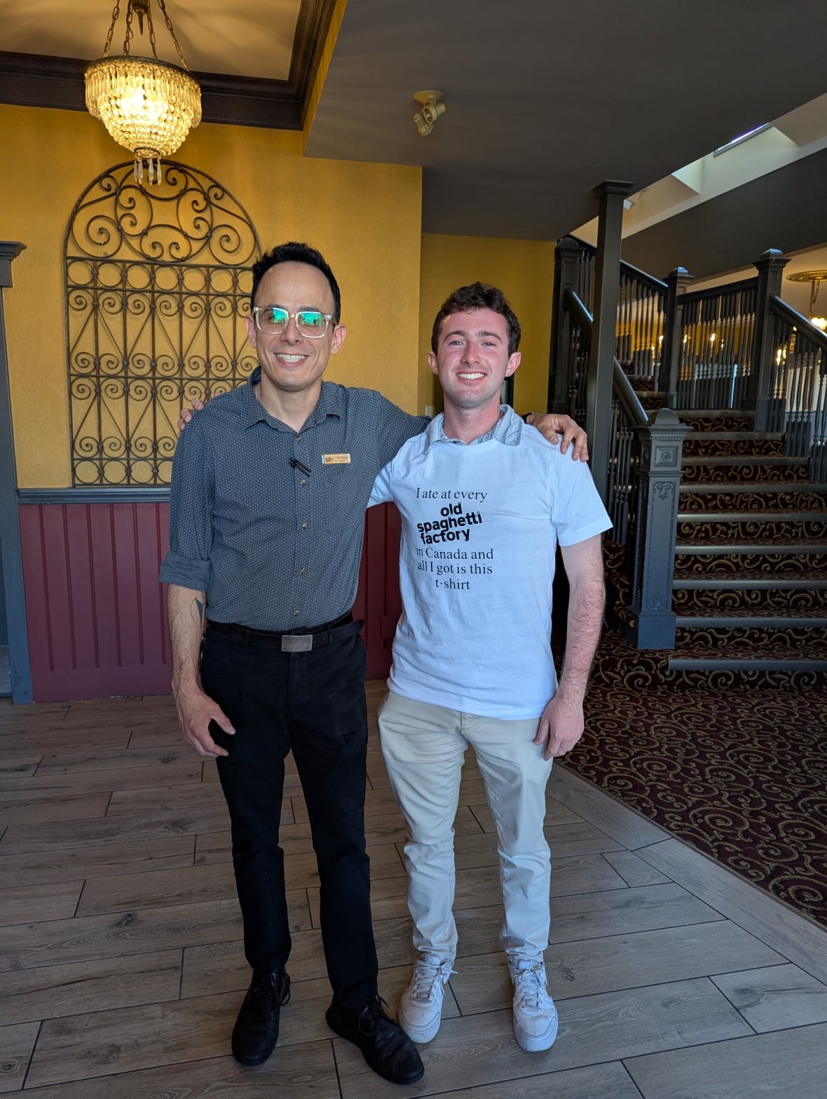 Two men stand next to each other: to the left, wearing grey shirt and black pants, to the right, wearing a custom Old Spaghetti Factory white shirt and khaki pants.