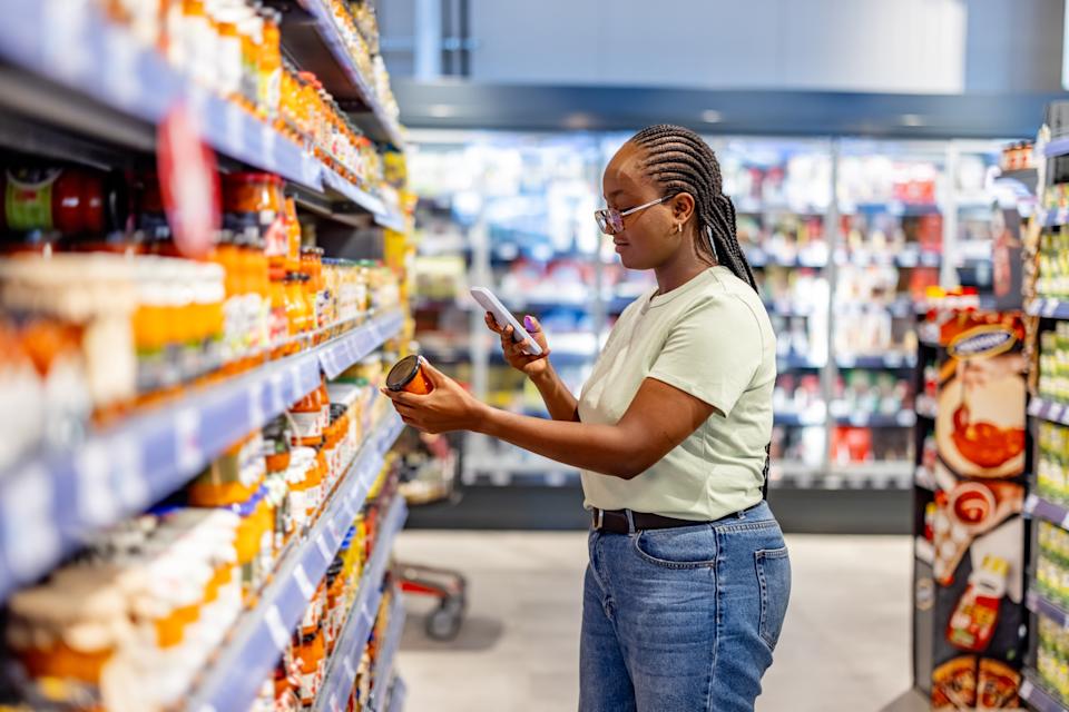A woman in casual attire shopping at a grocery store uses her smartphone to scan a jar for details. Shelves filled with packaged goods lend a bright and vibrant retail atmosphere.