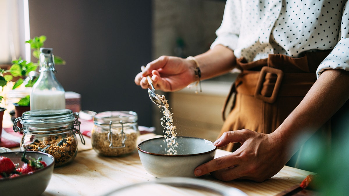 A woman in a sleeved polka dot shirt and apron prepares a healthy cereal breakfast in a sunlit kitchen, surrounded by fresh ingredients.