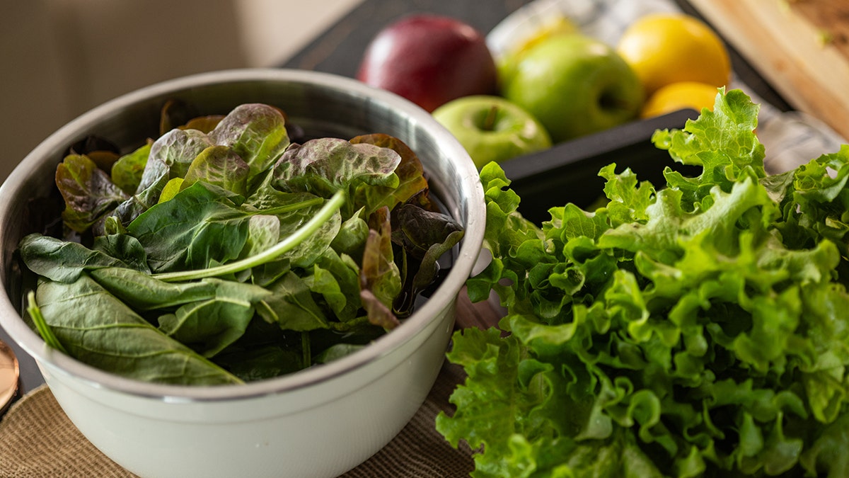 Fibrous leafy greens in a bowl next to more leafy greens, apples in the background.
