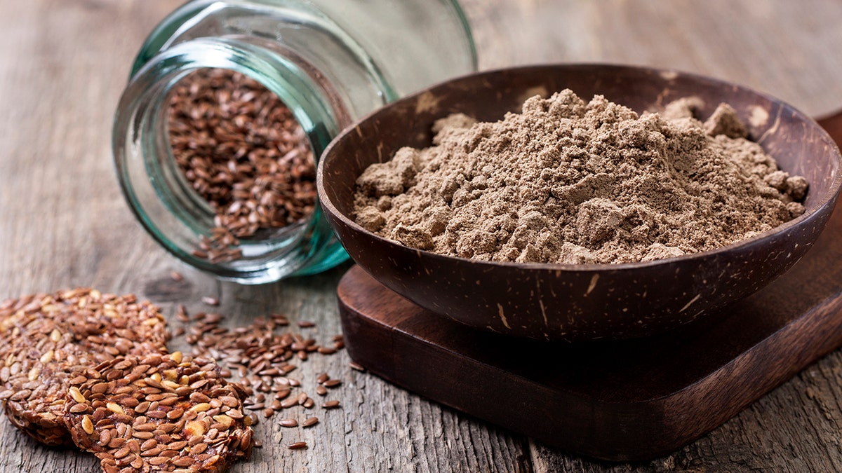 Ground up flax seeds pictured as powder in a wood bowl. In the background, a glass jar containing whole flax seeds is tilted on its side, spilling seeds.
