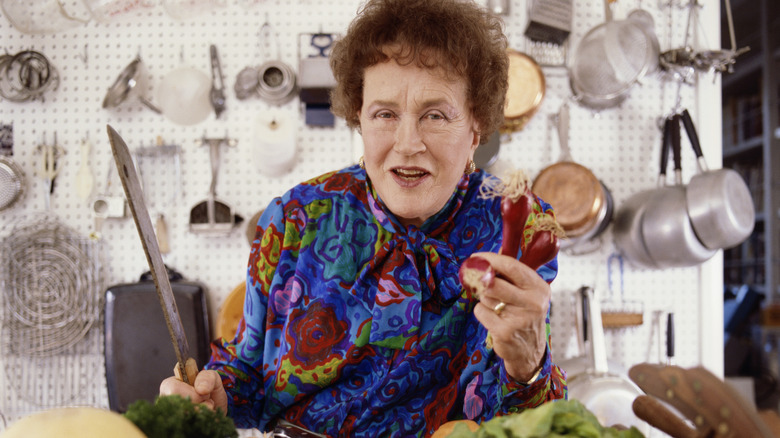 Julia Child poses in front of hanging kitchen tools