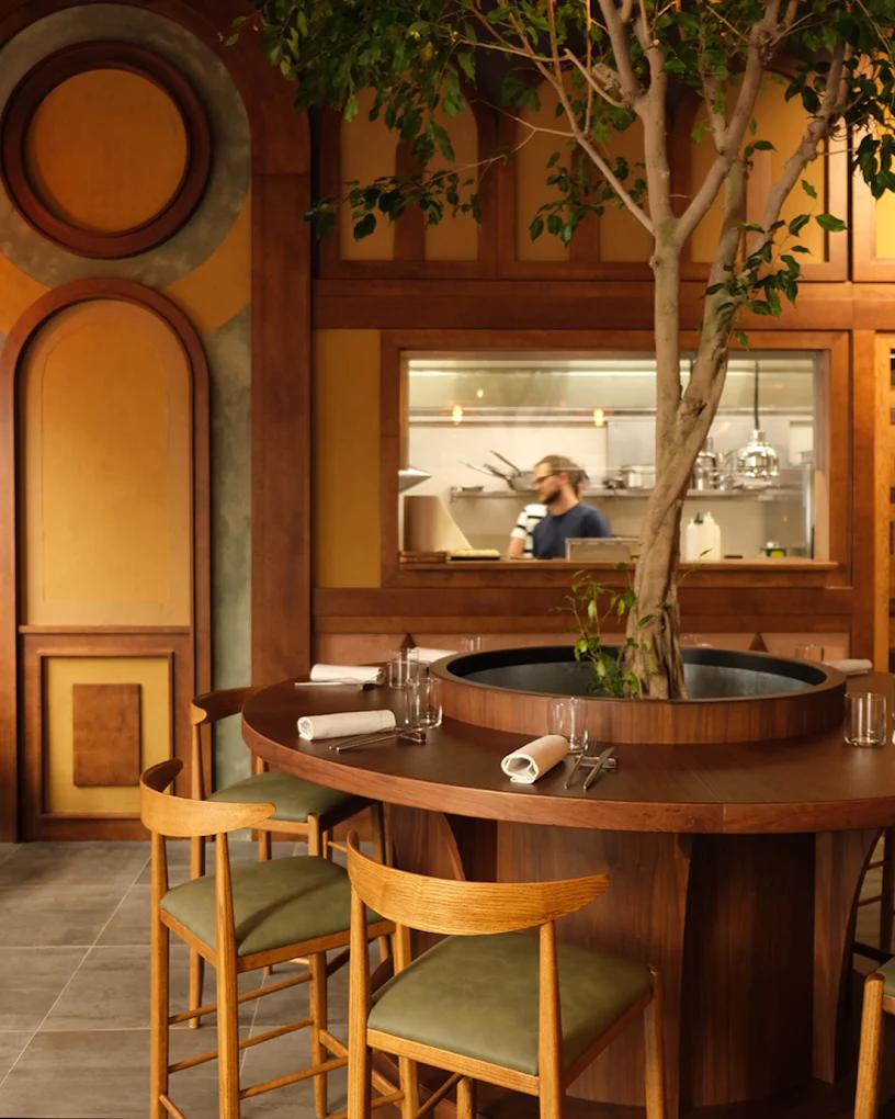 A warmly decorated Latin America-inspired restaurant features earthy, organic brown, green, and ochre tones, arched wall detailing, round wooden tables decorated with trailing plants sitting at their center, and wood and velvet chairs.