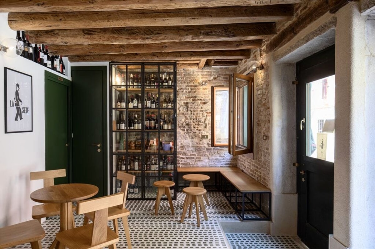 A quirkily designed wine bar features geometric floors in black and white tassels, sculptural wooden furniture, distressed brick walls, exposed beams, and a dark green, white, and brown color palette.