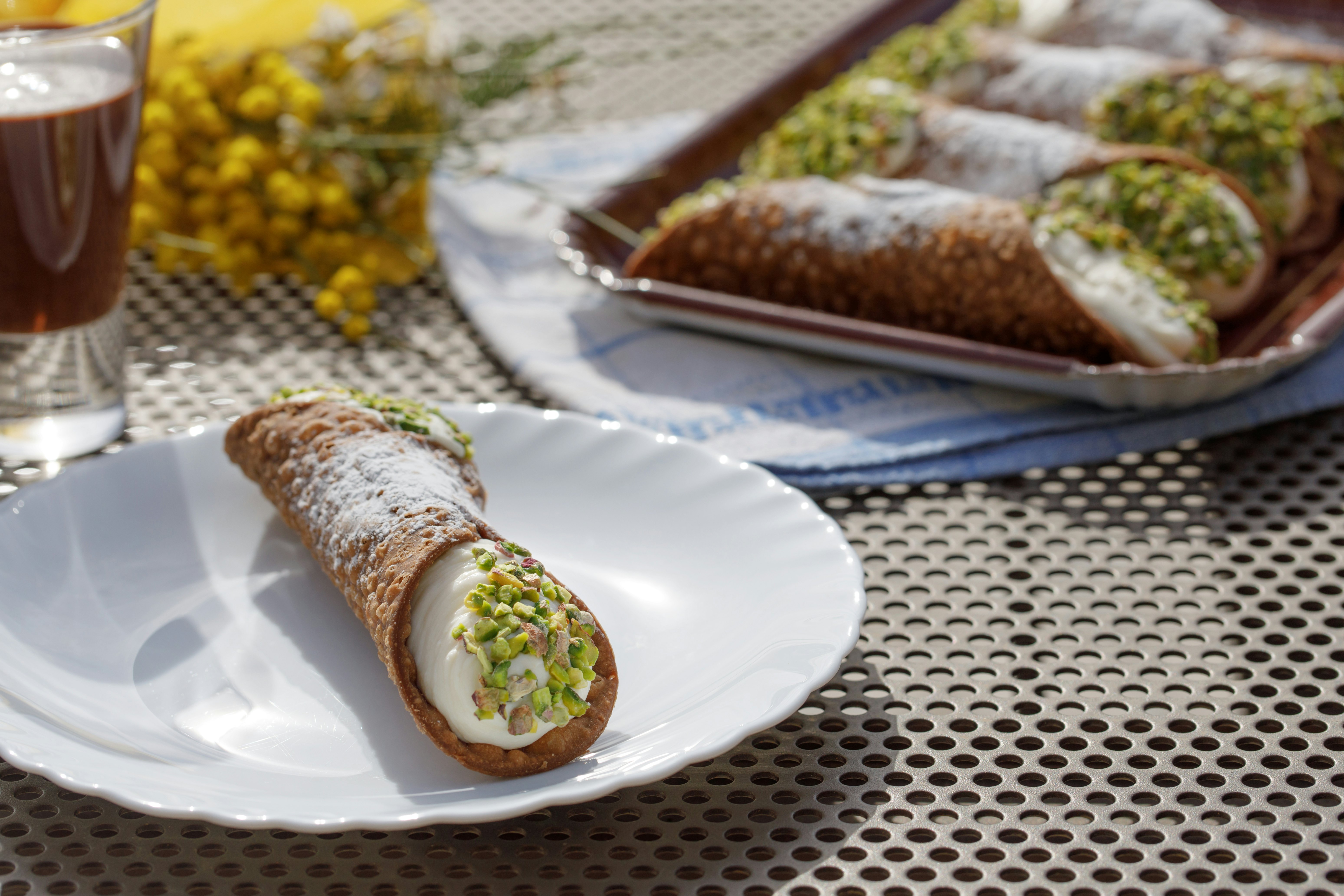 A white plate with one long pastry shell filled with cream, with green pistachios on the ends; more pastries are on a tray in the background.