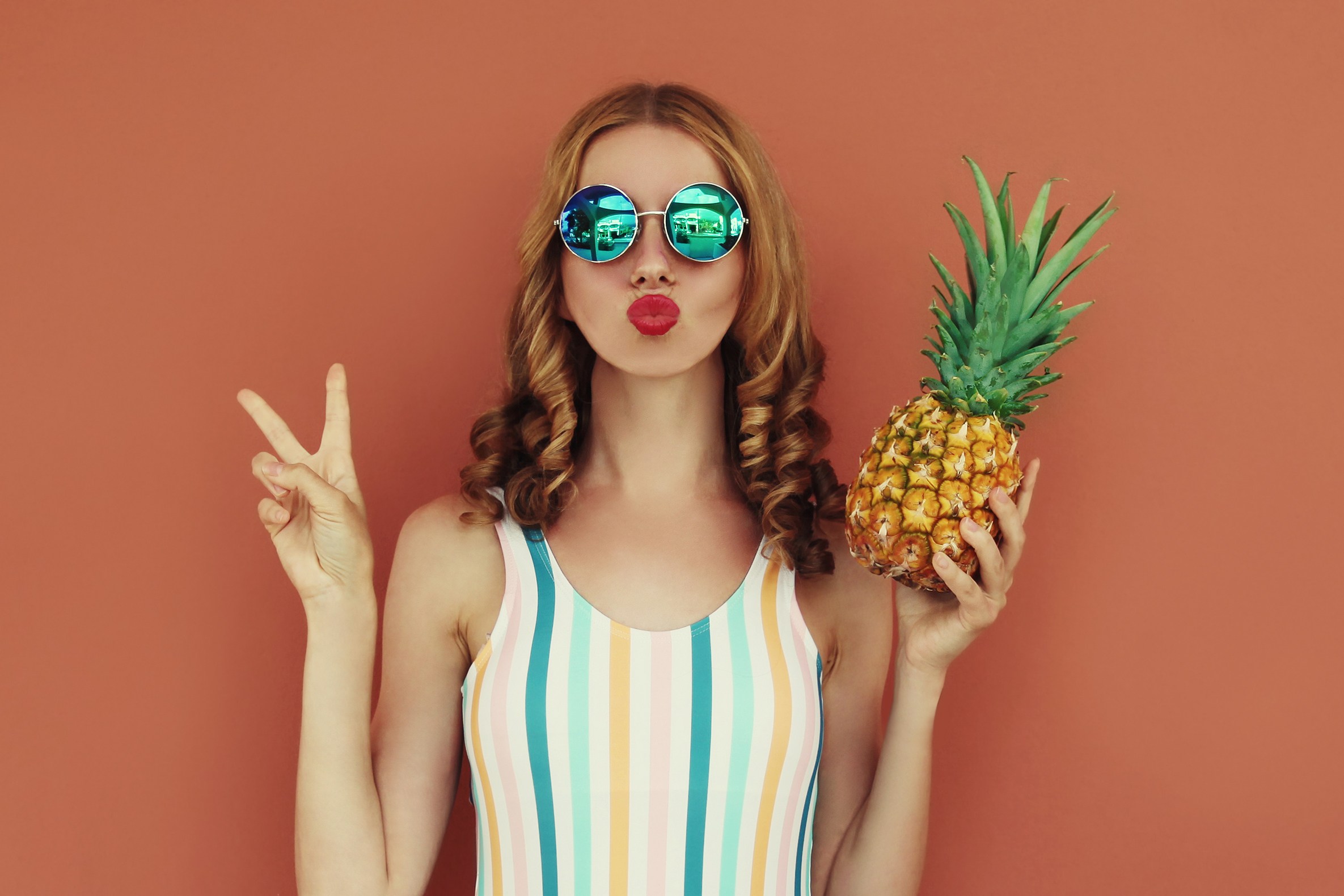 Woman in swimsuit and sunglasses holding a pineapple and giving the peace sign.