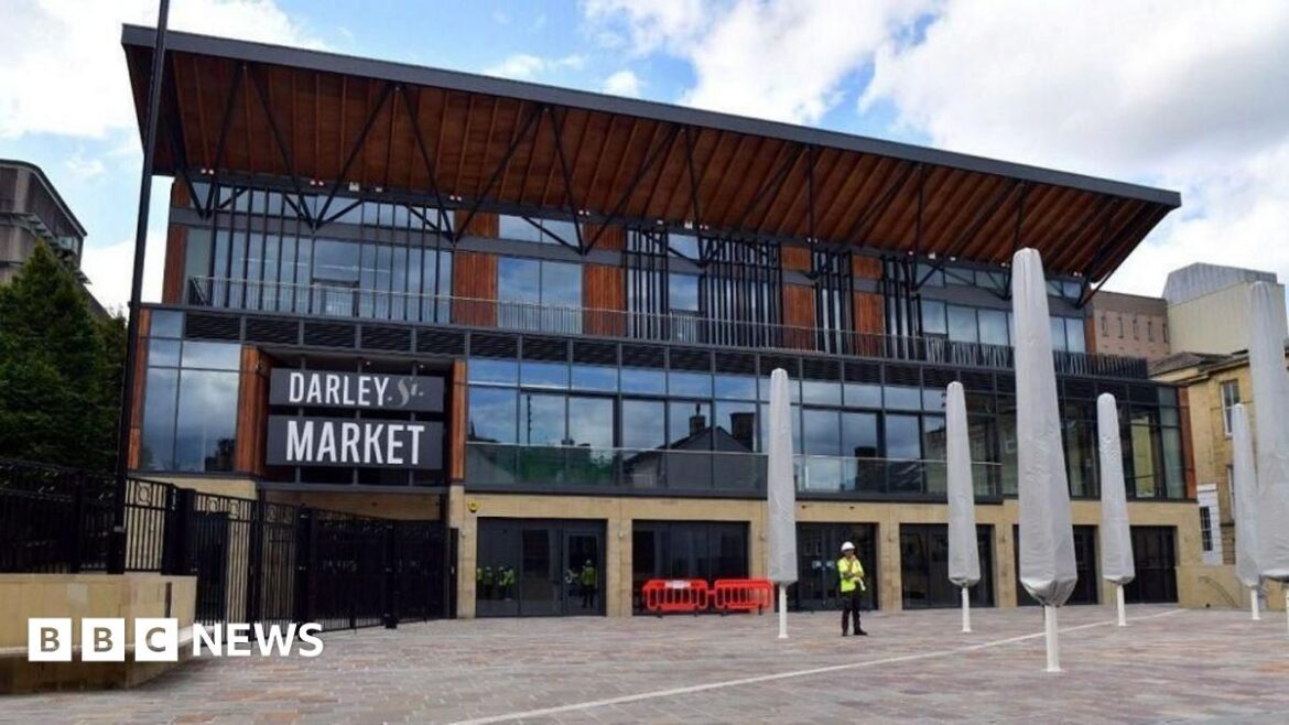Darley Street Market building is the focus of the photo. It has stone bottom, middle layer of glass and then a wooden roof. A man in a high viz stands in front of it.