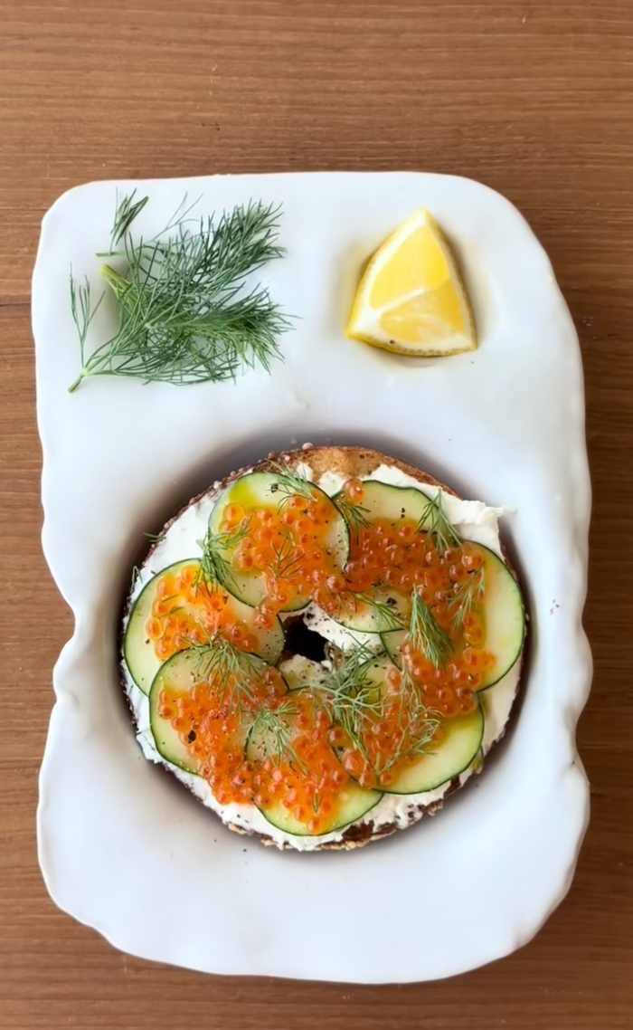 Bagel topped with cream cheese, cucumber slices, and salmon roe, garnished with dill. Lemon wedge and dill sprig on the side