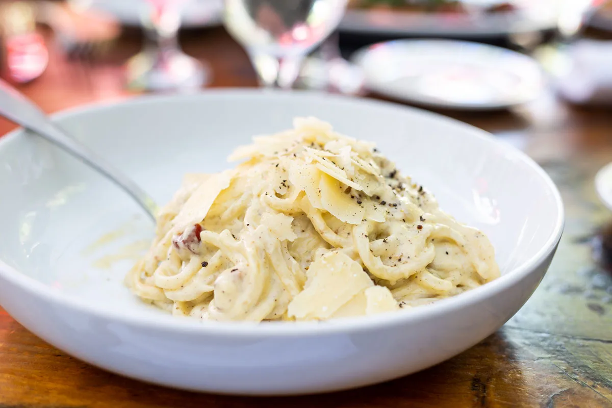 A white bowl of Fettuccine Alfredo with large flakes of parmesan cheese, and thick noodles, covered in sauce