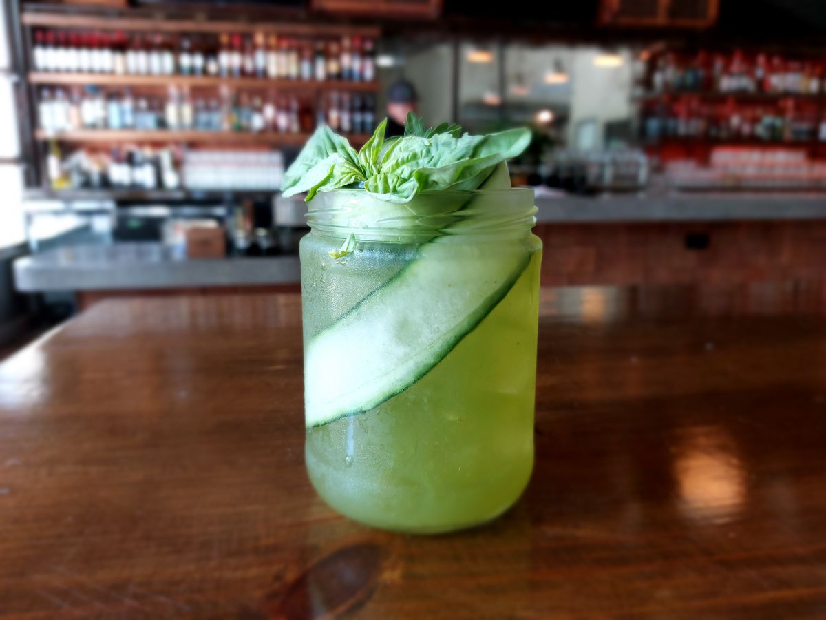 A green cocktail with a long cucumber slice in a jar on a brown table.