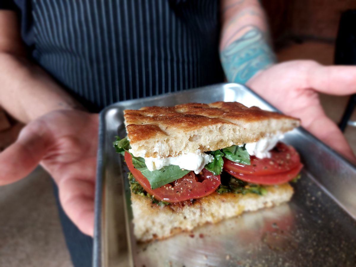 A focaccia sandwich with tomato slices, white cheese, and green leaves on a metal tray.