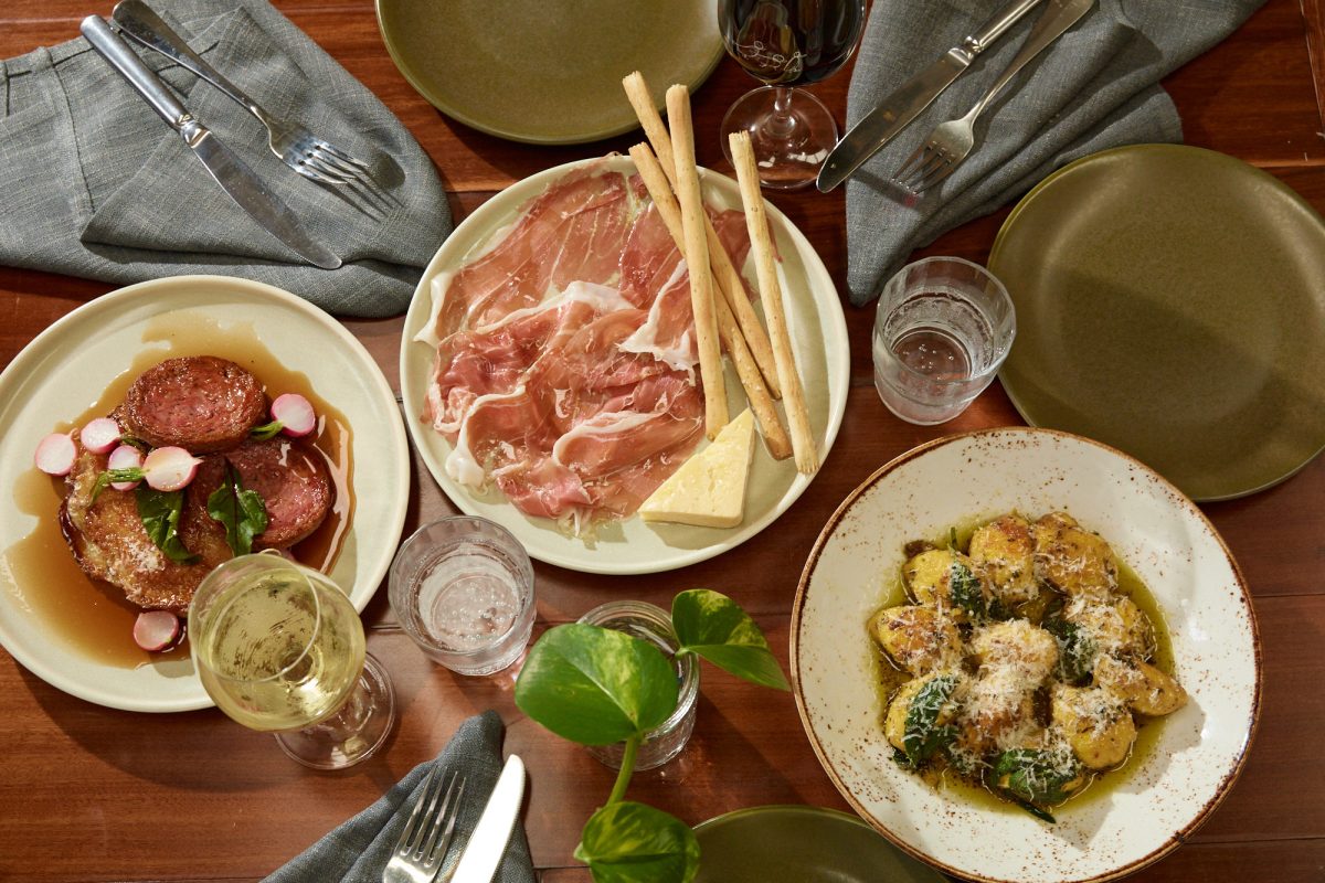 A table with three plates of food, one with sausages and radish, one with sliced cured meats, cheese and breadsticks and another with gnocchi. 