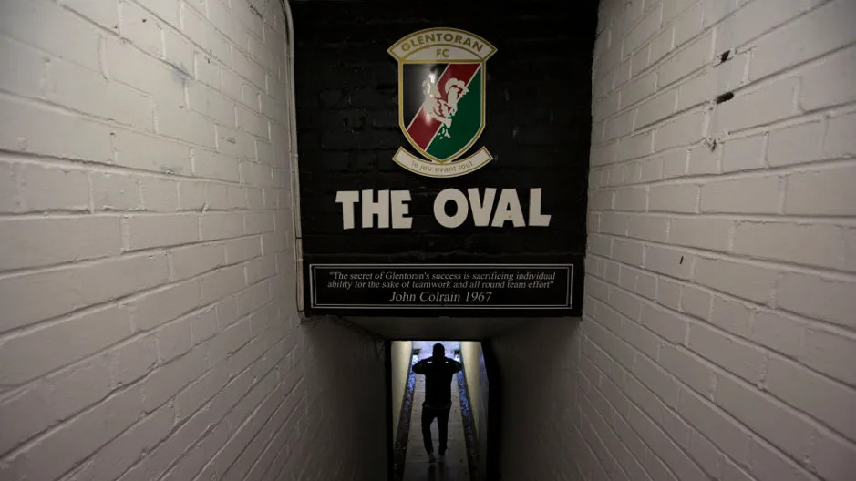 Man walks under an image saying The Oval, with a quote from former manager John Colrain underneath. There are two white walls on either side.