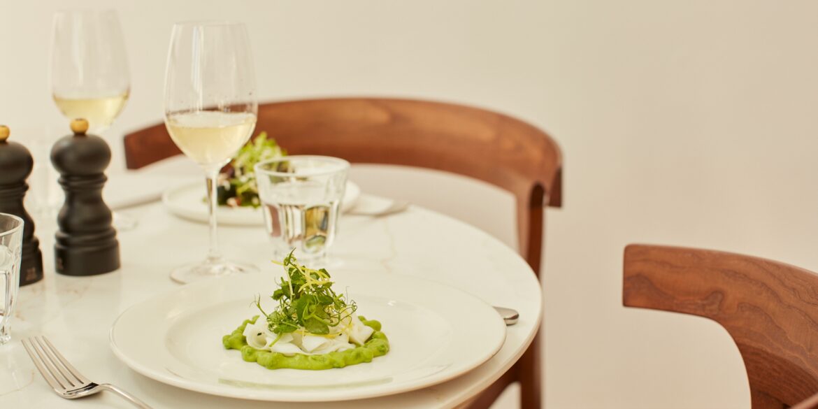 A restaurant table in marble and wood is complete with a white plate filled with a creamy broad beans and cheese starter, two white wine glasses, silver cutlery, and a salt and pepper grinder.