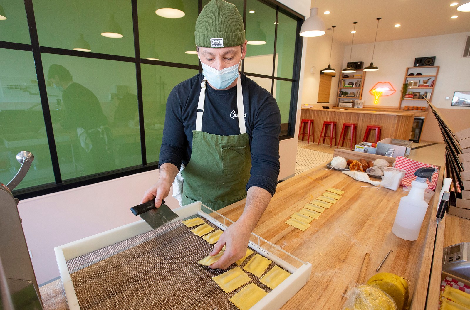 A man in a facemask and a beanie lays individual ravioli on a tray.