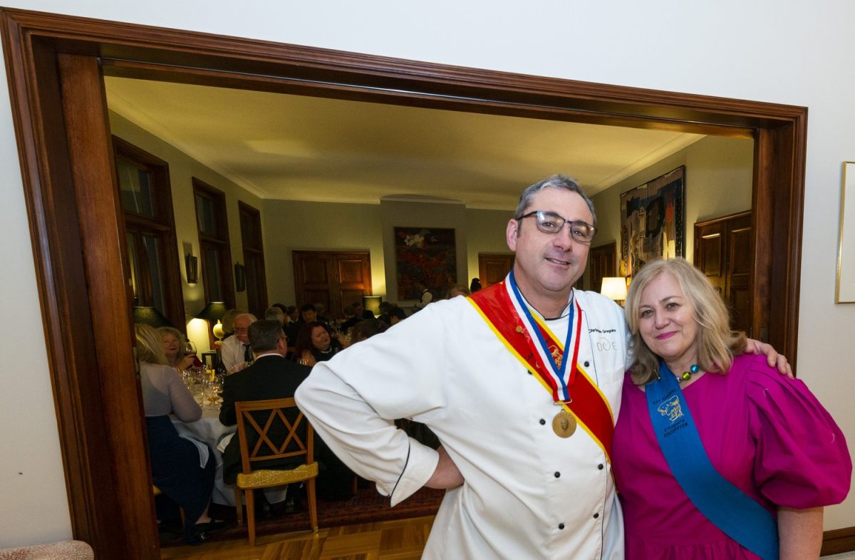A man (wearing medals) stands beside a woman in a pink dress and blue sash